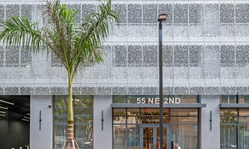 Street-level entrance of CMPND Miami at 55 NE 2nd Avenue with a perforated metal facade, palm tree, and glass lobby doors in downtown Miami