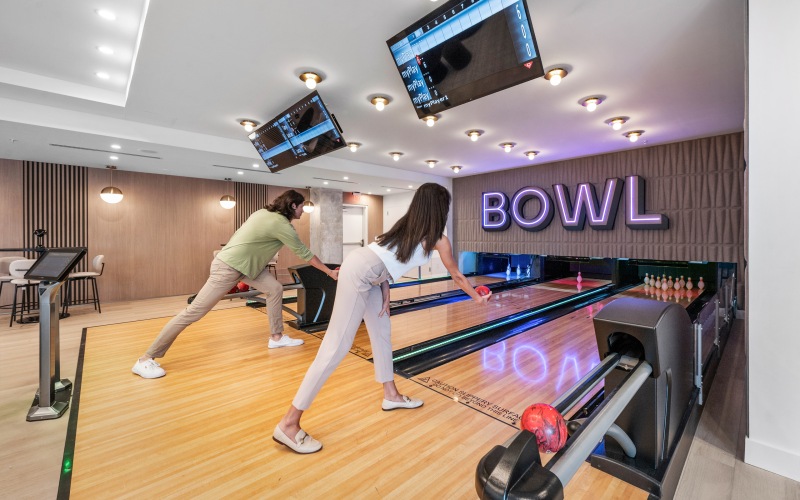 Man and woman bowling on the private indoor bowling lanes at CMPND Miami with a neon "BOWL" sign and overhead scoring screens