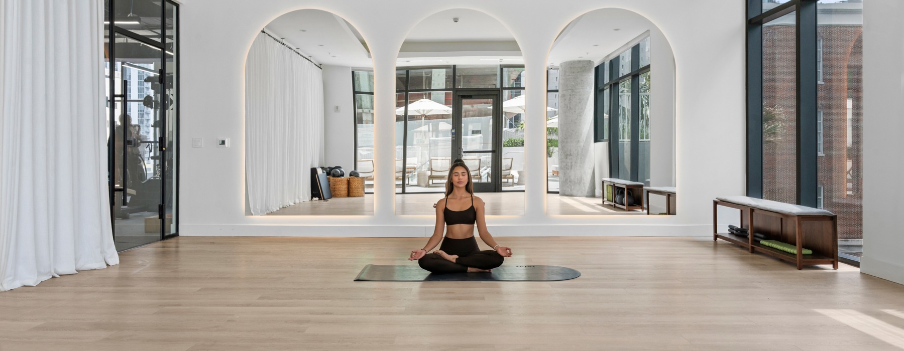 Woman meditating in a lotus pose on a mat in the CMPND Miami yoga studio with arched mirror alcoves and natural light